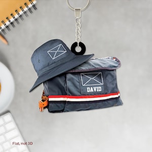 Könnte beinhalten: Eine blaue Canvas-Posttasche mit einem weißen Briefumschlag-Design und dem Namen "DAVID" darauf gedruckt. Die Tasche ist an einem blauen Bucket-Hut mit einem weißen Briefumschlag-Design befestigt. Hut und Tasche hängen an einem silbernen Schlüsselanhänger mit einem schwarzen Ring.