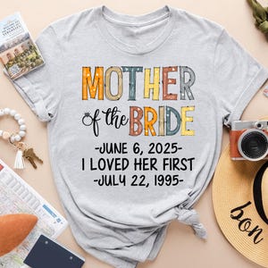 May include: A light grey t-shirt with the words "MOTHER of the BRIDE" in colourful letters. Below the text are the dates "JUNE 6, 2025" and "JULY 22, 1995". A camera, a hat, and a bracelet are visible in the background.