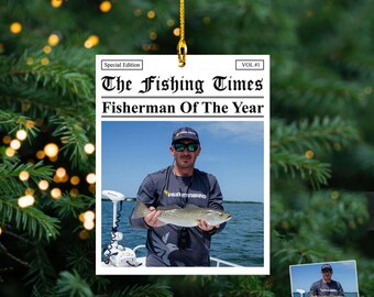 May include: A 2D acrylic ornament with a newspaper design. The headline reads "The Fishing Times Fisherman Of The Year." The ornament features a photo of a man holding a fish. The top of the ornament has a gold string for hanging.