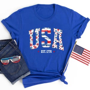 May include: A royal blue t-shirt with the word "USA" in a patriotic design, featuring floral and star patterns. The text "EST. 1776" is below the word "USA". The image also includes denim shorts, sunglasses, and a small American flag.