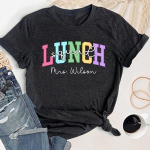 Puede incluir: Camiseta gris oscuro con las palabras "LUNCH squad" en letras de bloque coloridas y el nombre "Mrs. Wilson" en cursiva. La camiseta está anudada en la parte inferior y combinada con jeans azules y una taza de café.