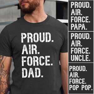 May include: Dark grey t-shirt with the words "PROUD. AIR. FORCE. DAD." in white, distressed lettering. Additional text on the right side reads "PROUD. AIR. FORCE. PAPA.", "PROUD. AIR. FORCE. UNCLE.", and "PROUD. AIR. FORCE. POP POP."