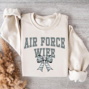 May include: A cream-coloured sweatshirt featuring the text "AIR FORCE WIFE" in grey block letters. A grey camouflage bow is positioned below the text. The sweatshirt is folded on a light-coloured surface.