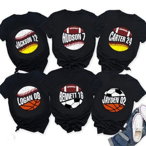 May include: Six black t-shirts with sports-themed designs. Each shirt features a different name and number inside a graphic of a baseball, football, basketball, or soccer ball. The names and numbers are in white.