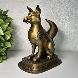 Puede incluir: Estatua de zorro de color bronce con detalles intrincados, sentada sobre una base rectangular. El zorro tiene orejas puntiagudas, una cola rizada y patrones decorativos en su cuerpo. La estatua se exhibe sobre una superficie blanca.