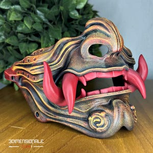Può includere: Una dettagliata maschera da demone giapponese stampata in 3D nei toni del marrone, dell'oro e del rosso. La maschera presenta prominenti zanne rosse e una lunga lingua rossa. La maschera ha dettagli intricati ed è progettata con un'espressione feroce.