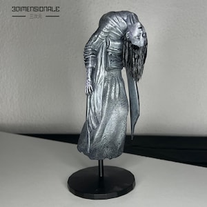May include: A silver-colored figurine of a woman with long hair, wearing a flowing gown, standing on a black base. The figurine is titled "3DIMENSIONAL E."
