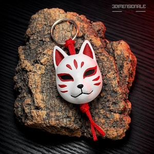 May include: A white fox mask keychain with red accents and a black nose and mouth. The mask is attached to a silver keyring with a red cord. The keychain is displayed on a piece of textured, brown bark.