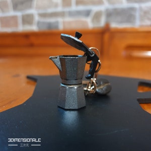 May include: A silver metal keychain shaped like a small coffee pot with a black lid that is open. The keychain is on a black surface.