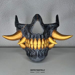 Puede incluir: Una máscara de calavera negra y dorada con dientes y cuernos afilados. La máscara está hecha de un material texturizado y tiene un diseño detallado.