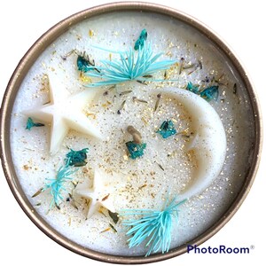 May include: A white candle with a gold glitter finish and embedded white stars and a crescent moon. The candle is decorated with dried flowers and blue pine needles.