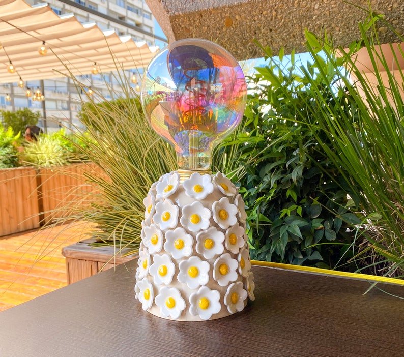 May include: A white ceramic lamp base with a cluster of white flowers with yellow centers. The lamp has a clear glass bulb with a rainbow iridescent finish.