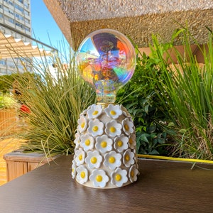 May include: A white ceramic lamp base with a floral design featuring white daisies with yellow centers. The lamp base is topped with a clear iridescent glass globe lightbulb.