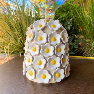 May include: A ceramic lamp base with a white glaze and yellow flower accents. The base is shaped like a bell and is covered in small, raised flowers.