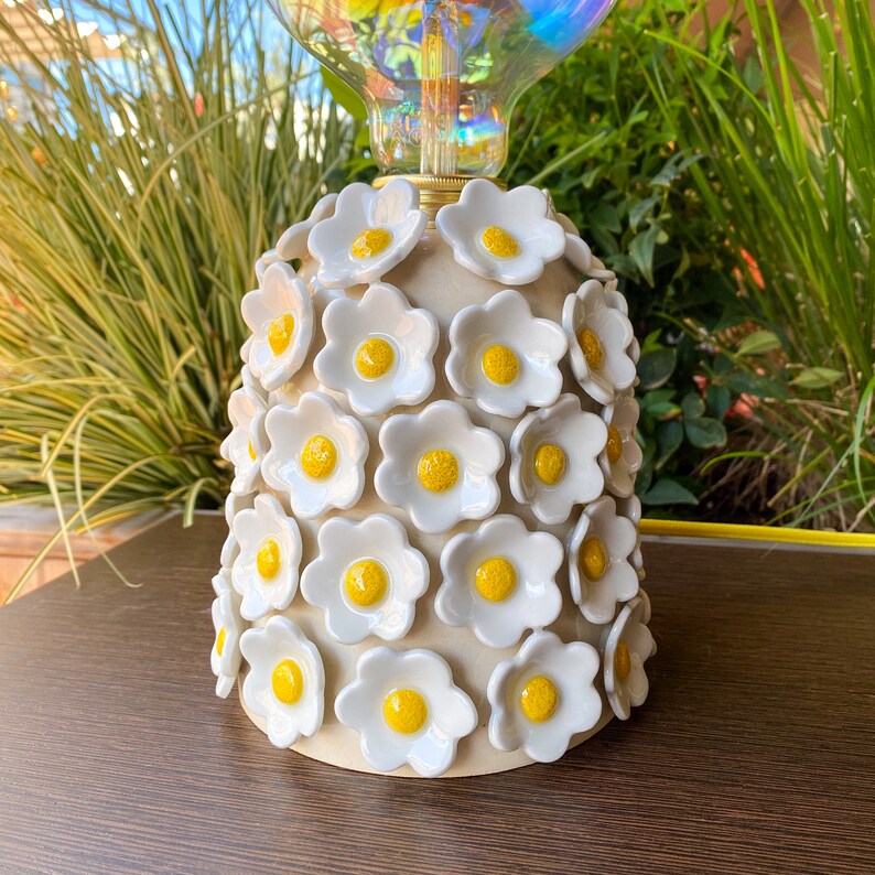 May include: A ceramic lamp base with a white glaze and yellow flower accents. The base is shaped like a bell and is covered in small, raised flowers.