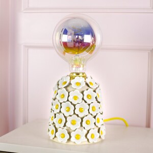 May include: A white ceramic table lamp with a floral design. The lamp has a clear glass bulb with a rainbow iridescent finish. The lamp is on a white surface.