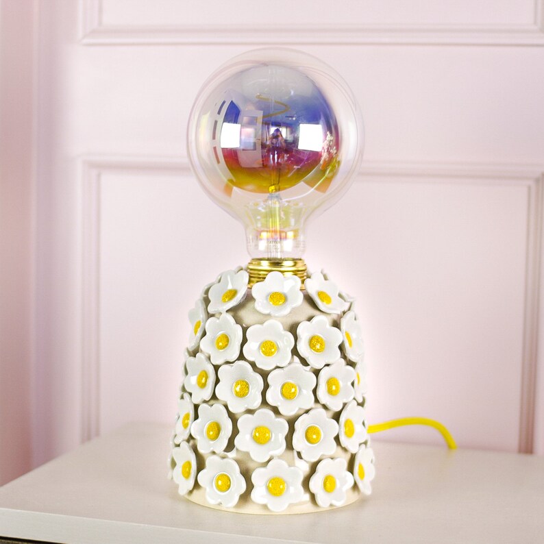May include: A white ceramic table lamp with a floral design. The lamp has a clear glass bulb with a rainbow iridescent finish. The lamp is on a white surface.