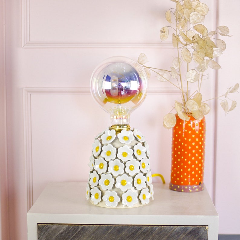 May include: A white ceramic table lamp with a clear glass globe shade and a base decorated with white daisies with yellow centers. The lamp is on a white nightstand with a vase of dried flowers.