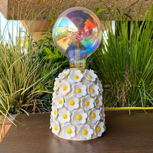May include: A white ceramic lamp base with a cluster of white and yellow daisy-shaped flowers. The lamp is topped with a clear glass globe with iridescent rainbow colors.