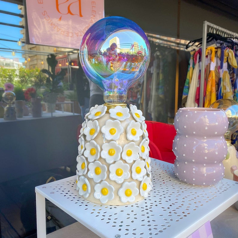 May include: A white ceramic lamp with a floral design. The lamp has a clear glass globe with a rainbow iridescent finish. The lamp is sitting on a white table with a decorative pattern.