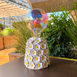 May include: A white ceramic lamp base with a floral design featuring yellow centers. The lamp base is topped with a clear iridescent light bulb.
