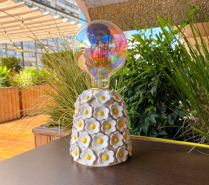 May include: A white ceramic lamp base with a floral design featuring yellow centers. The lamp base is topped with a clear iridescent light bulb.