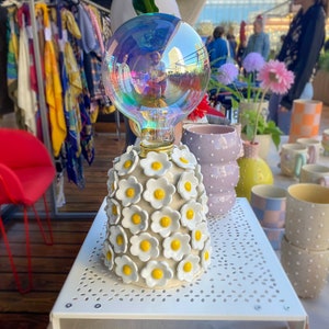 May include: A white ceramic lamp base with a white and yellow daisy design. The lamp base is topped with a clear glass globe with a rainbow iridescent finish. The lamp is sitting on a white table with a white and gray patterned surface.