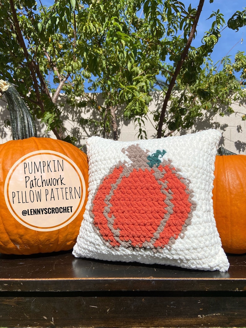 K&ouml;nnte beinhalten: Ein wei&szlig;es H&auml;kelkissen mit einem Patchwork-K&uuml;rbisdesign in Orange, Braun und Gr&uuml;n. Das Kissen liegt auf einer Holzoberfl&auml;che mit zwei echten K&uuml;rbissen auf beiden Seiten. Der Text "Pumpkin Patchwork Pillow Pattern @Lennyscrochet" befindet sich auf dem K&uuml;rbis links.