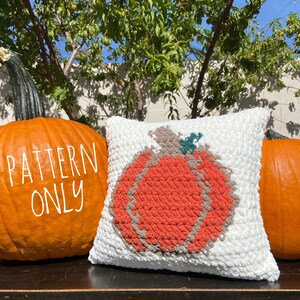 K&ouml;nnte beinhalten: Ein wei&szlig;es geh&auml;keltes Kissen mit einem gro&szlig;en orangefarbenen K&uuml;rbisdesign. Das Kissen liegt auf einer Holzoberfl&auml;che mit zwei echten K&uuml;rbissen auf beiden Seiten. Der Text "PATTERN ONLY" ist auf den K&uuml;rbis links geschrieben.