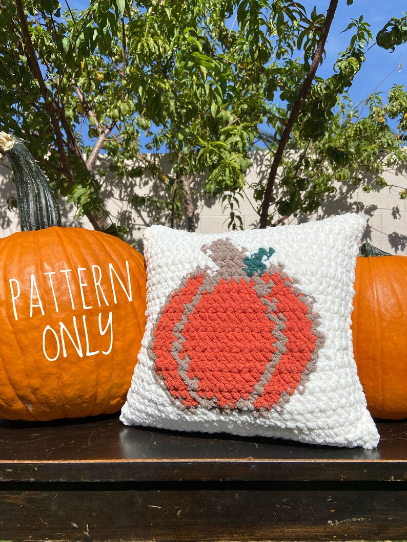 K&ouml;nnte beinhalten: Ein wei&szlig;es geh&auml;keltes Kissen mit einem gro&szlig;en orangefarbenen K&uuml;rbisdesign. Das Kissen liegt auf einer Holzoberfl&auml;che mit zwei echten K&uuml;rbissen auf beiden Seiten. Der Text "PATTERN ONLY" ist auf den K&uuml;rbis links geschrieben.