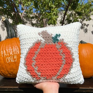 K&ouml;nnte beinhalten: Ein wei&szlig;es H&auml;kelkissen mit einem gro&szlig;en orangefarbenen K&uuml;rbisdesign. Der K&uuml;rbis hat einen braunen Stiel und gr&uuml;ne Bl&auml;tter. Das Kissen liegt auf einem Holztisch mit zwei K&uuml;rbissen im Hintergrund.