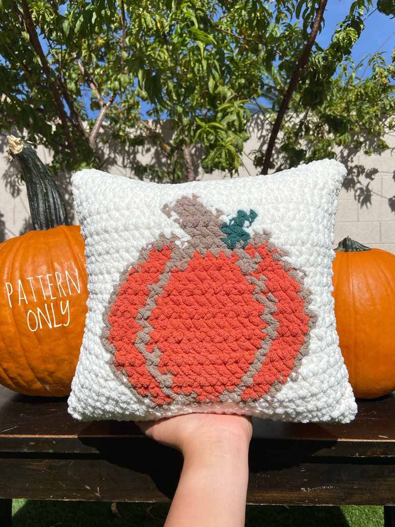 K&ouml;nnte beinhalten: Ein wei&szlig;es H&auml;kelkissen mit einem gro&szlig;en orangefarbenen K&uuml;rbisdesign. Der K&uuml;rbis hat einen braunen Stiel und gr&uuml;ne Bl&auml;tter. Das Kissen liegt auf einem Holztisch mit zwei K&uuml;rbissen im Hintergrund.