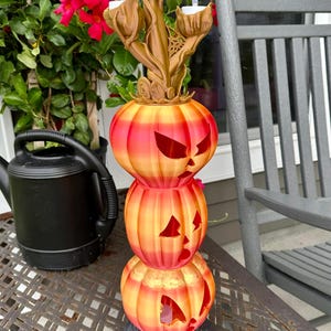 Pode incluir: Um porta-velas de abóbora de três níveis com um caule dourado e três porta-velas. As abóboras são laranja e vermelho com um efeito degradado. O porta-velas está sobre uma mesa com um padrão de grelha metálica.