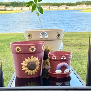 May include: Three terracotta flower pots with painted sunflower and rainbow designs. The largest pot is brown with yellow sunflowers, the middle pot is red with a large sunflower, and the smallest pot is red with a rainbow and clouds.
