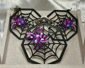 May include: Three black spider web-shaped keychains with a purple and blue spider in the center of each web. The keychains are displayed in a white box with a decorative border.