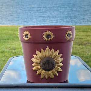 May include: A burgundy-colored flower pot with a large sunflower design on the front and smaller sunflowers around the rim. The pot is made of a solid material and is suitable for planting flowers or herbs.