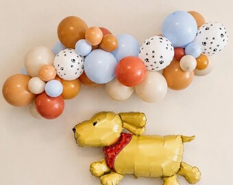 May include: A balloon garland featuring a mix of round balloons in shades of tan, brown, rust orange, light blue, and white with black paw prints, accompanied by a single gold foil balloon shaped like a dog wearing a red patterned bandana.