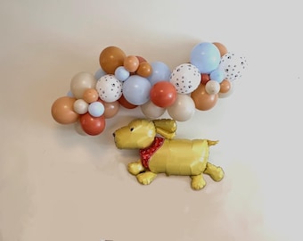 May include: A balloon arch made of light blue, beige, brown, and white balloons with a black paw print design. A yellow dog balloon is in the foreground.