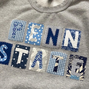 May include: A gray sweatshirt with the words "PENN STATE" appliqued across the front. The letters are in a variety of blue and white patterned fabrics, including polka dots, stripes, and gingham. The sweatshirt is a casual, comfortable style.