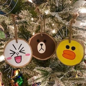 May include: Three wooden Christmas ornaments hanging on a tree. Each ornament is a circular wood slice with a cartoon face painted on it. One is white with a laughing face, one is brown with a bear face, and one is yellow with a duck face.