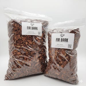 May include: Two clear plastic bags filled with brown fir bark. Each bag has a white label with the text "CRITTERS CAVE FIR BARK" and a QR code. The bags are sealed at the top and are likely for use in terrariums or as a substrate for reptiles.