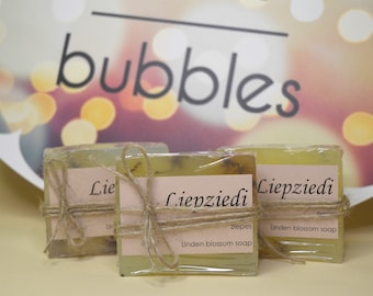Puede incluir: Tres pastillas de jabón de flor de tilo, envueltas en plástico transparente y atadas con hilo de bramante. Cada pastilla tiene una etiqueta que dice "Liepziedi" y "Linden blossom soap". El fondo presenta la palabra "bubbles" y luces bokeh.