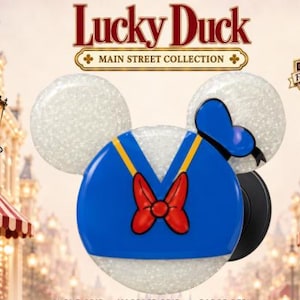 May include: A circular phone grip featuring a blue sailor shirt design with a red bow, yellow trim, and a blue hat. The accessory has two white glittery ears and is labeled Lucky Duck Main Street Collection, Fan Inspired, and Grandma's Main Street Designs.
