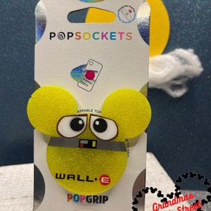 May include: A yellow and black Wall-E PopGrip phone accessory with a glitter finish. The PopGrip is shaped like Wall-E's head and has a black strap across the middle. The text "WALL-E" is printed on the bottom of the PopGrip. The PopGrip is packaged on a white card with the text "POPGRIP" and "APPABLE TOP" printed on it. The card also has a black and white Mickey Mouse logo with the text "Grandmas Main Street Designs" printed on it.