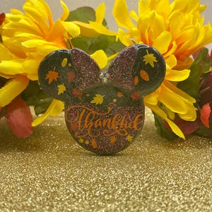 May include: A black and glittery Minnie Mouse-shaped ornament with a purple bow, decorated with fall leaves. The word "Thankful" is written in orange script across the front. The ornament is set against a gold glitter background with yellow flowers.