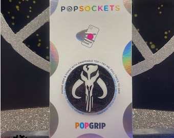 Può includere: Un PopSocket per telefono nero e bianco con un simbolo mandaloriano. Il PopSocket è su una carta bianca con le parole "PopSockets" e "PopGrip" stampate su di essa.