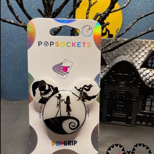 May include: A white and black phone grip with a silhouette of Jack Skellington and Sally from the Nightmare Before Christmas. The grip is shaped like Mickey Mouse ears and has the words "PopGrip" and "PopSockets" on the packaging.