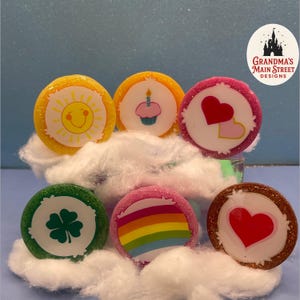 May include: A collection of colorful, glittery round designs featuring various graphics, including a smiling sun, a cupcake, hearts, a four-leaf clover, a rainbow, and a single heart. The designs are set against a blue background with cotton-like material.