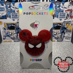 Könnte beinhalten: Roter und schwarzer Spider-Man PopGrip Handygriff mit Glitzer-Finish. Der Griff hat ein Spinnennetz-Design und eine schwarze Spinne auf der einen Seite und eine rote Spider-Man-Maske auf der anderen Seite. Die Verpackung enthält den Text "PopSockets" und "PopGrip".