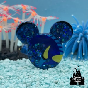 May include: A blue glittery Mickey Mouse-shaped pin featuring a Dory design from Finding Nemo. The pin is set against a backdrop of blue and orange coral and light blue pebbles. The pin has a yellow fin and is surrounded by blue glitter.